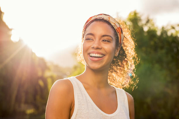 Young african woman smiling at sunset Portrait of beautiful african american woman smiling and looking away at park during sunset. Outdoor portrait of a smiling black girl. Happy cheerful girl laughing at park with colored hair band. happiness stock pictures, royalty-free photos & images