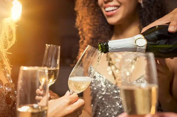 Hand pouring champagne from bottle into glasses with friends around him. Closeup of hand pouring white wine in flutes during party. Detail shot of new year"u2019s eve celebration.