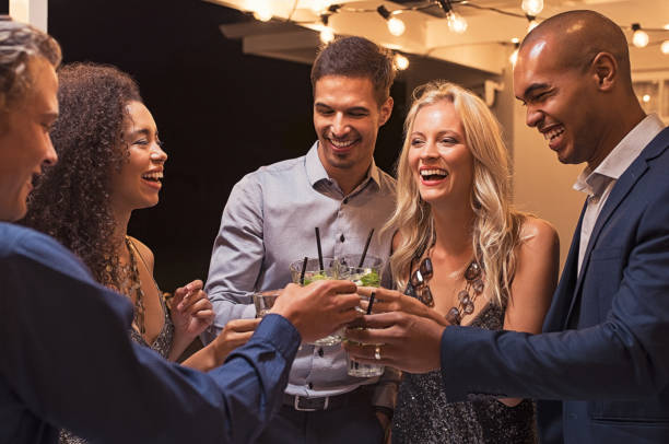 Friends toasting cocktail at night party Group of young multiethnic friends enjoying evening and drinking cocktails. Happy men and women raising a toast with mojito on a patio under the light bulb wire. Elegant girls and stylish guy having fun together at party night. cocktail party photos stock pictures, royalty-free photos & images