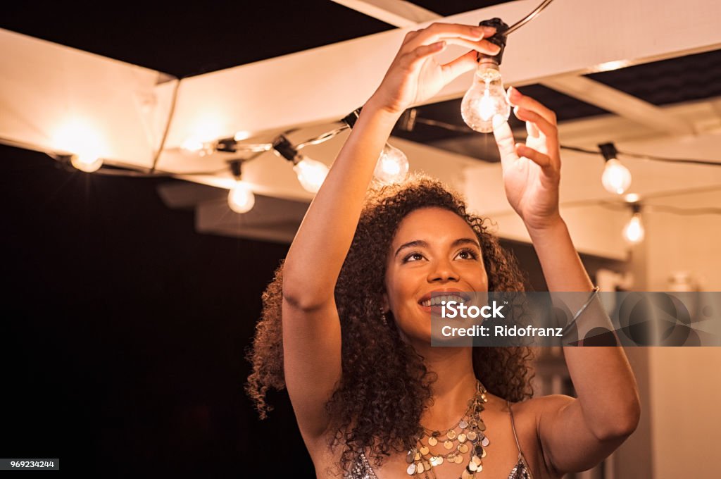 Woman setting up for party Cheerful young woman wearing party dress changing bulb light in patio. Happy smiling girl making preparation of party by adding lights outdoor. Happy beautiful woman enjoying fixing bulbs in backyard. Lighting Equipment Stock Photo