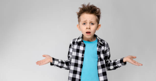 A handsome boy in a plaid shirt, blue shirt and jeans stands on a gray background. The boy spread his hands in both directions Charming happy child on gray background. The boy's hair is up. The boy has a hairstyle. The boy spread his hands in both directions confused face stock pictures, royalty-free photos & images