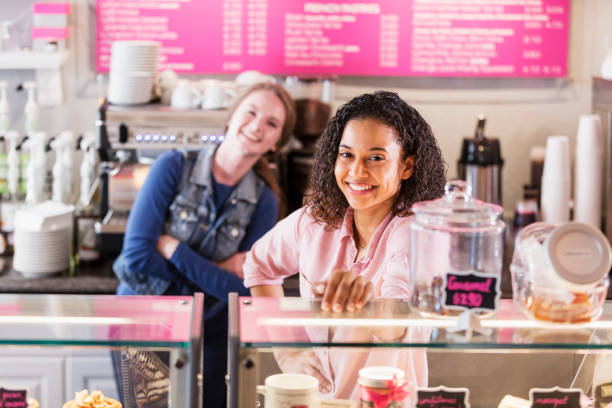 Two multi-ethnic young women in local pastry shop Two young multi-ethnic women working in a local coffee or pastry shop standing behind the display case, smiling at the camera. The focus is on the mixed race African-American and Caucasian woman in the foreground. franchising photos stock pictures, royalty-free photos & images