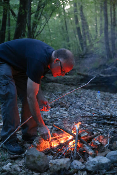 istock Man Tending Building Camping Campfire Near Stream at Twilight 969302176