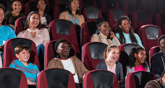 A large multi-ethnic group of children, teens and young adults sitting in a movie theater, watching a movie. It must be a comedy as the audience is laughing.