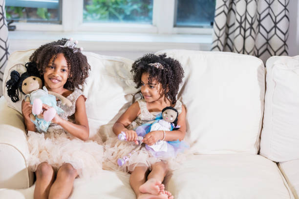 Mixed race sisters playing with dolls Two cute mixed race African-American and Hispanic girls, sisters 3 and 5 years old, playing together, sitting side by side on a sofa, holding dolls in their laps, wearing princess dresses and tiaras, smiling at the camera. girl playing with doll stock pictures, royalty-free photos & images