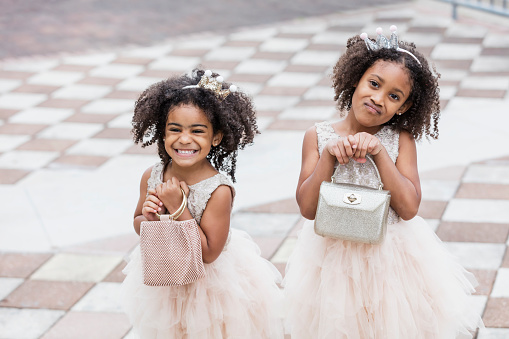 Two mixed race African-American and Hispanic girls, sisters 3 and 5 years old, wearing dresses and tiaras, holding purses. They are smiling and grinning at the camera.