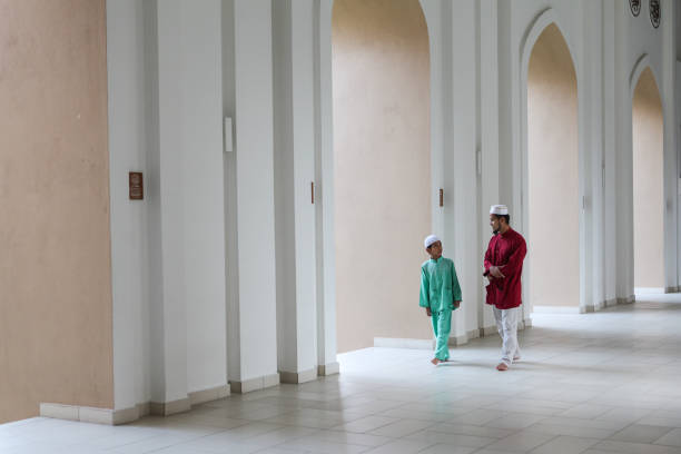 A Malay Muslim father and his son walking together in a mosque, wearing a traditional Malay dress for men, heading towards the prayer hall.