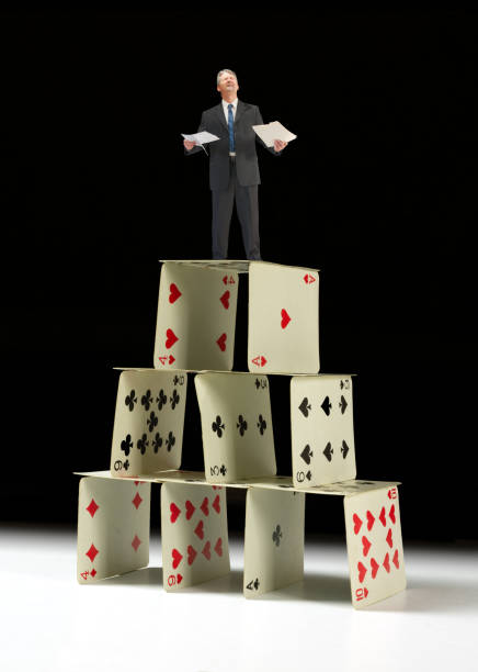 Man in business suit standing on house of cards Distraught man in business suit holding work folders standing on house of cards representing financial difficulties, work stress, crashing stock market, housing bubble, ponzi scheme, fraud, etc. ponzi scheme stock pictures, royalty-free photos & images