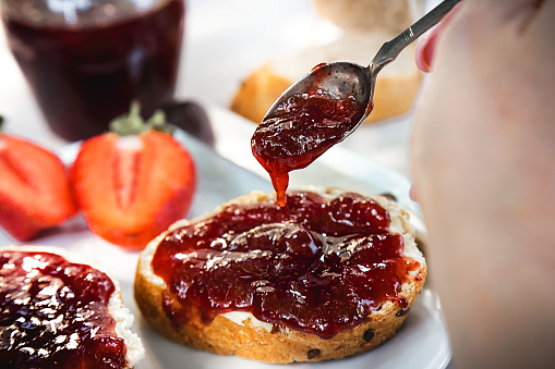Hand with spoon pouring homemade organic strawberry jam over slices of bread with butter  on a white plate at wooden table. Served with glass of fresh milk.