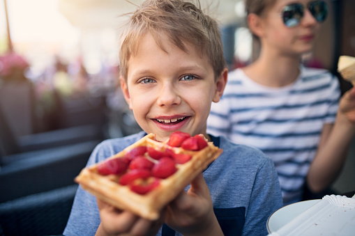 Little boy aged 8 eating waffle with strawberries. Sister eating ice cream in the background.\nNikon D850.