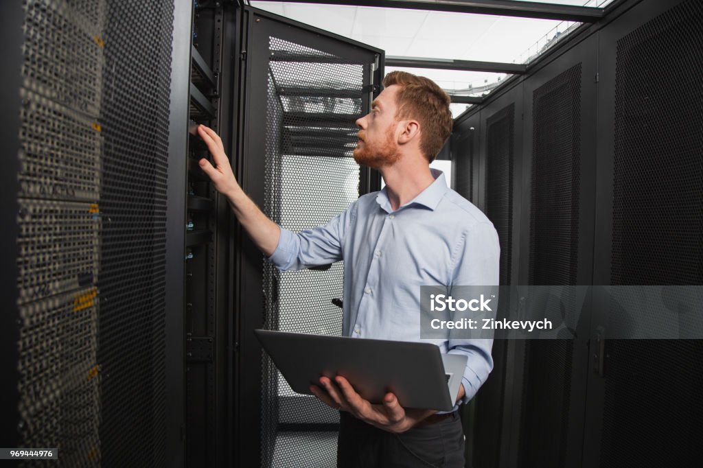 Skillful IT technician establishing connection Computer programming. Confident IT technician examining server closet while carrying laptop Accessibility Stock Photo