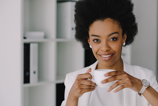 attractive african american businesswoman holding cup of coffee and looking at camera in office