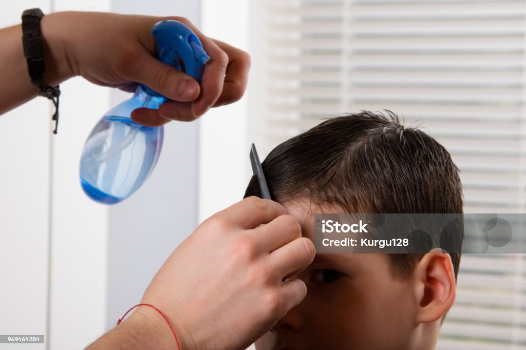 the hairdresser to the boy moisturizes his hair with a spray of water and makes a hairdo Adult Stock Photo