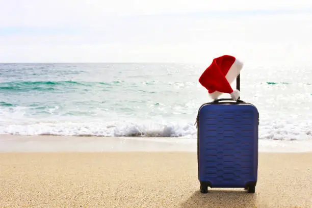 Photo of One hard shell suitcase on a beach. Tropical destination vacation concept. Santa claus hat on extended telescoping luggage handle.