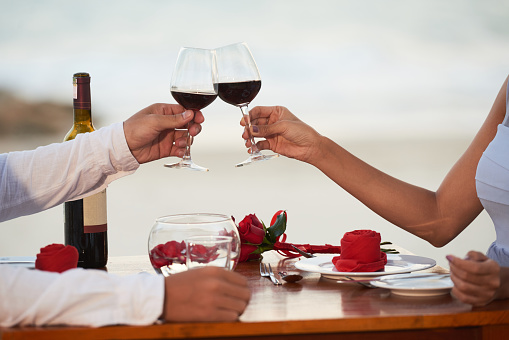 Close-up shot of young couple sitting at table of beach restaurant and clinking wine glasses together while celebrating momentous event, blurred background