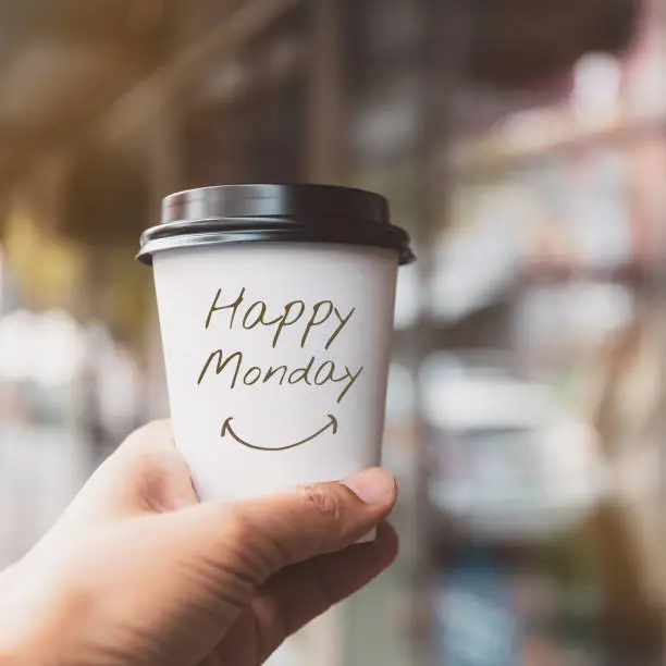 Hand holding white coffee paper cup with text "HAPPY MONDAY" on blurred window background.Vintage tone, Encouragement and Motivation concept.