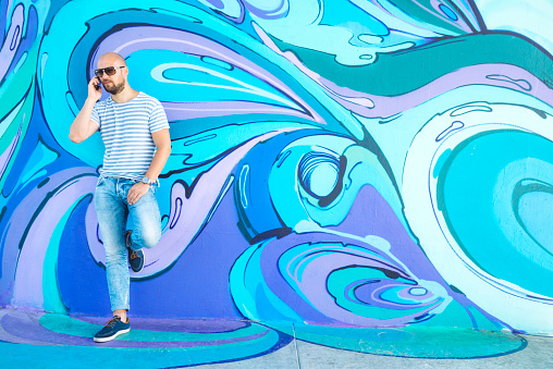 Hipster male with sunglasses and mobile phone next to wall of colorful  graffiti in Belgrade