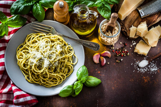 Spaghetti al pesto Genoese plate shot from above on dark kitchen table. Ingredients for preparing pesto sauce like olive oil, pine nuts, basil leaves, salt, pepper, garlic and Parmesan cheese are around the plate. Low key DSRL studio photo taken with Canon EOS 5D Mk II and Canon EF 100mm f/2.8L Macro IS USM