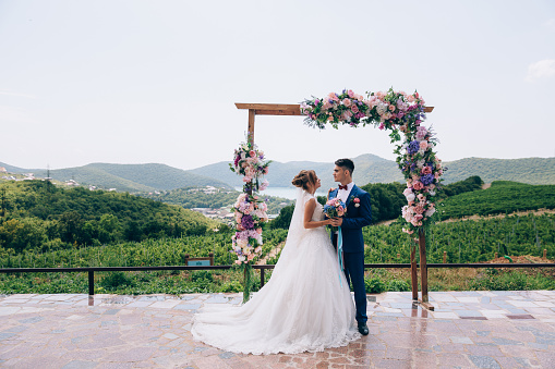 Newlyweds in love look at each other and enjoy the wedding day. They stand on an arch of pink, white and blue flowers