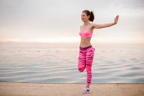 Smiling young woman dressed in pink sportswear doing morning exercise near the seaside during sunrise
