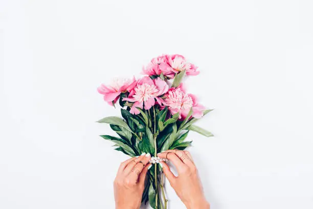 Women's hands decorate a bouquet of pink peony flowers with  pearls, top view. Flat lay minimal composition with copy space.