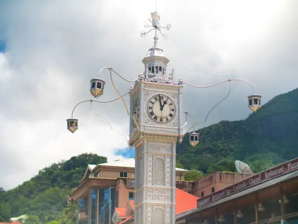 Victoria Clock Tower in Victoria, Seychelles. Bright but cloudy day with green jungle covered hills in background.