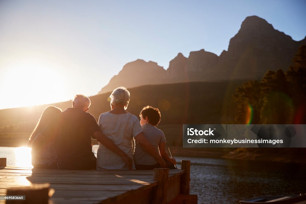 Grandchildren With Grandparents Sitting On Wooden Jetty By Lake Family Stock Photo