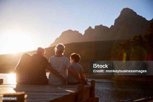 Grandchildren With Grandparents Sitting On Wooden Jetty By Lake Stock Photo - Download Image Now