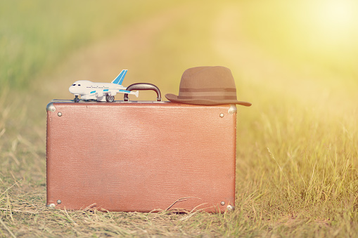 Travel and adventure concept. Vintage brown suitcase and hat with toy airplane near the road in the green field.