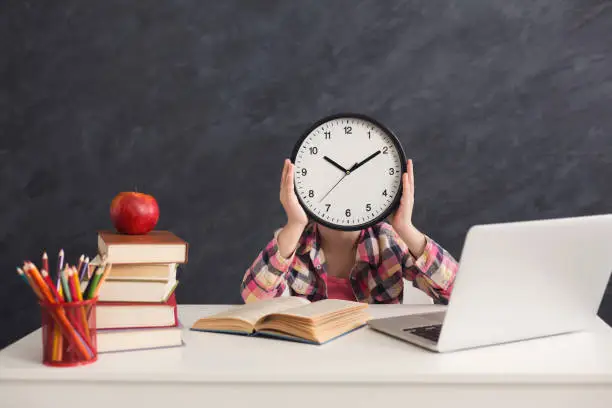 Smart girl sitting with stack of books and laptop, holding big clock, covering face. Education, development, time management, deadline, time to study, school concept