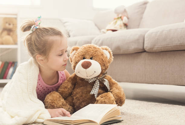 istock Happy little girl and her teddy bear reading book 969603636