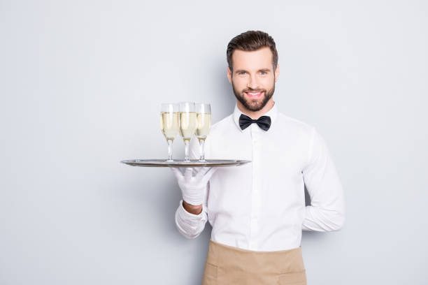 portret radosnego przyjaznego mężczyzny w białej klasycznej koszuli i czarnej kokardce trzymającej rękę za plecami i tacą z trzema kieliszkami wina musującego, odizolowanego na szarym tle, patrzącego na kamerę - butler champagne service waiter zdjęcia i obrazy z banku zdjęć