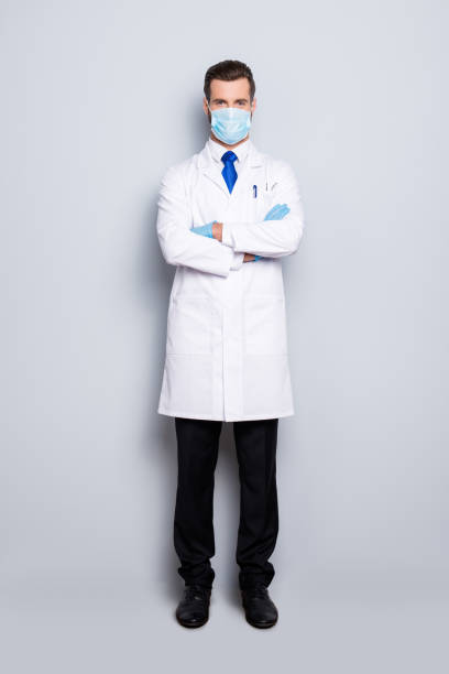 full size fullbody portrait of attractive handsome dentist with hairstyle in protective face mask, white lab coat, blue tie, having crossed hands, isolated on grey background - dentist dental hygiene dental assistant dentist office imagens e fotografias de stock