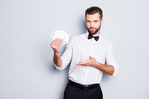 Portrait of stylish trendy croupier with hairstyle in formal wear having, showing with palm set of cards in hands, isolated on grey background, looking at camera