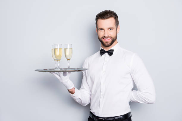 портрет веселого дружелюбного человека в классической белой рубашке и черном луке, держащем руку за спиной и подносом с тремя бокалами шам� - butler champagne service waiter стоковые фото и изображения