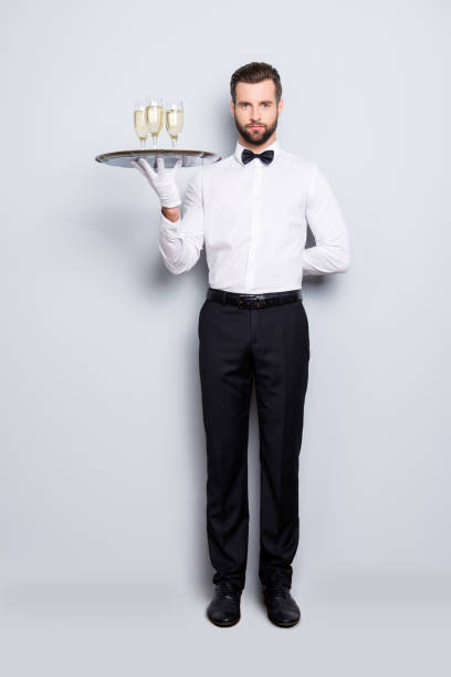 pełnowymiarowy portret pełnowymiarowego mężczyzny w klasycznej białej koszuli i czarnej muszce trzymającej rękę za plecami i tacą z trzema kieliszkami wina musującego, wyizolowanym na szarym tle - butler champagne service waiter zdjęcia i obrazy z banku zdjęć