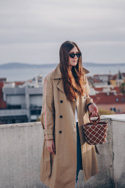 fashion blogger posando en una azotea en su elegante gabardina beige con una raya de lado, pantalones de mezclilla, zapatos de correa de tobillo, usar gafas de sol. la celebración de un cuero bolso de totalizador de la red. - vertical caucasian glasses red hair fotografías e imágenes de stock