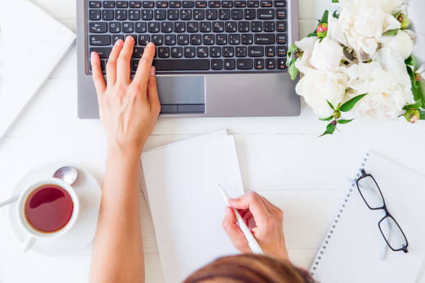 istock Workspace with girl's hand on laptop keyboard and writing in notebook with empty blank, cup of tea, glasses, white peony on woodden table. Top view feminine office desk. Freelancer working place. 969662436