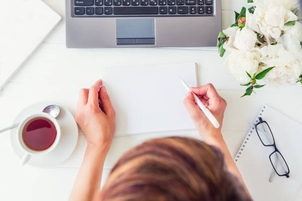 istock Workspace with girl's hands writing in notebook with empty blank, laptop keyboard, cup of tea, glasses, white peony on woodden table. Top view feminine office desk. Freelancer working place. 969662468