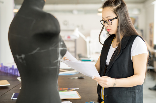 Side view portrait of focused Asian woman working in fashion design holding sketch standing at tailors table by sewing dummy in modern atelier studio