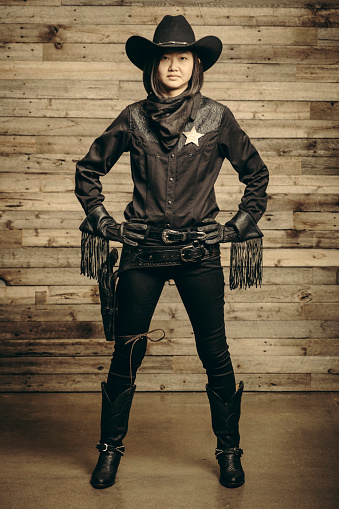 A Japanese woman dressed as a cowboy sheriff in front of an aged wooden wall.