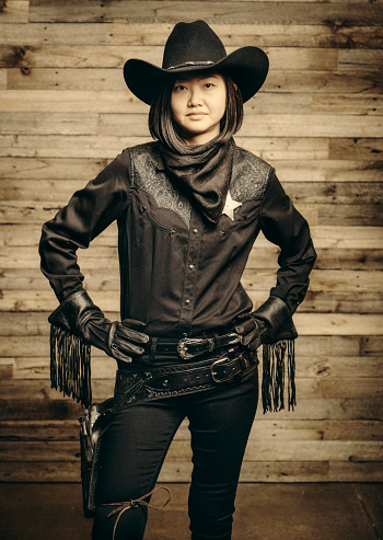 A Japanese woman dressed as a cowboy sheriff in front of an aged wooden wall.