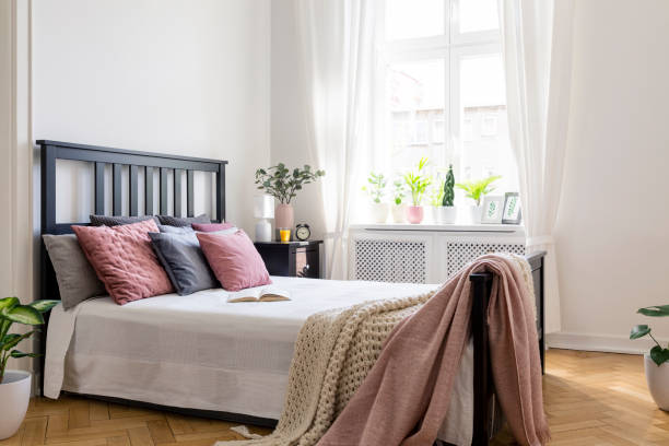 istock Pink blanket on bed with black headboard in bright bedroom interior with plants and window. Real photo 969910218
