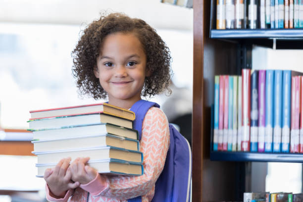 Young mixed race girl smiles while holding a large stack of library books. She is looking at the camera.