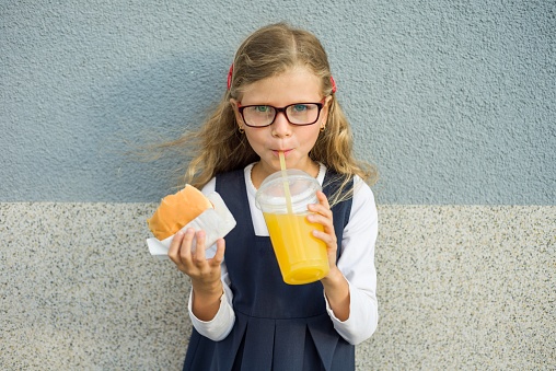 Outdoor portrait of child girl with blond curly hair with glasses. Girl eats sandwich and drinks orange juice. Background gray wall