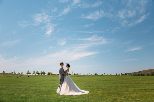 newlyweds are walking along the green field of the golf club on a wedding day. The groom in a business suit is gray and the bride in a luxury white dress with a veil are holding hands