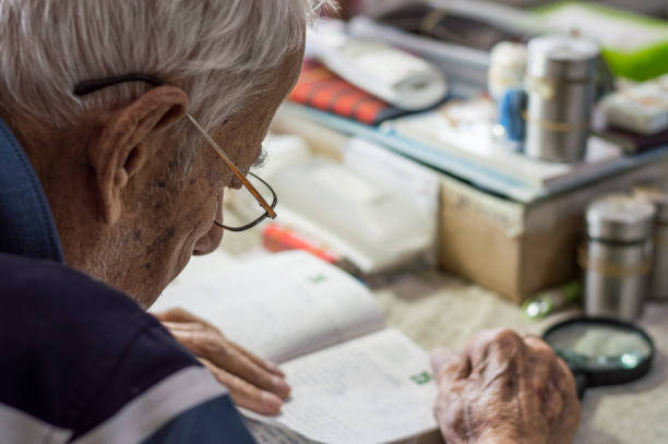 Elderly man with glasses reading writings in notebook near the window at home stock photo