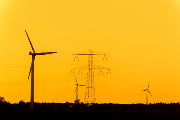 Overhead power lines in front of wind turbines and a power plant in the background during sunset.
