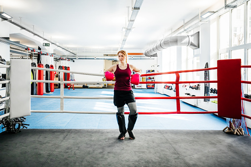 A portrait of a young, female, amateur kickboxer wearing protective gear and standing in the  ring at her local gym.