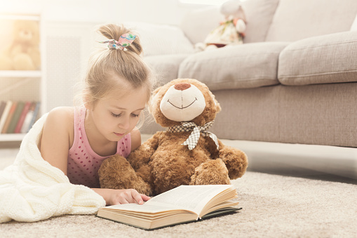 Cute happy little casual girl embracing teddy bear and reading book, wrapped in white blanket. Pretty kid at home, lying on floor near sofa with her favorite toy, copy space
